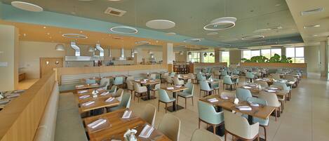 A spacious dining area with multiple tables and chairs, a kitchen in the background, and large windows providing natural light.