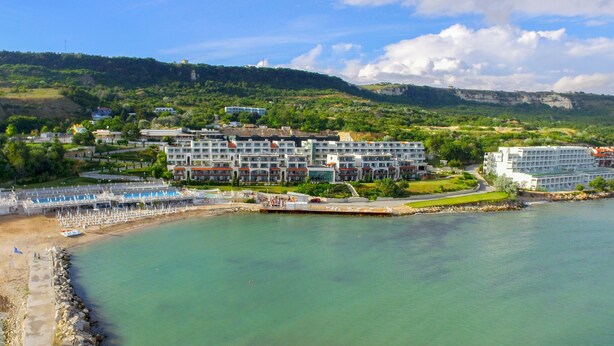 A coastal hotel with multiple buildings, a beach area with umbrellas, and a view of the sea and hills in the background.