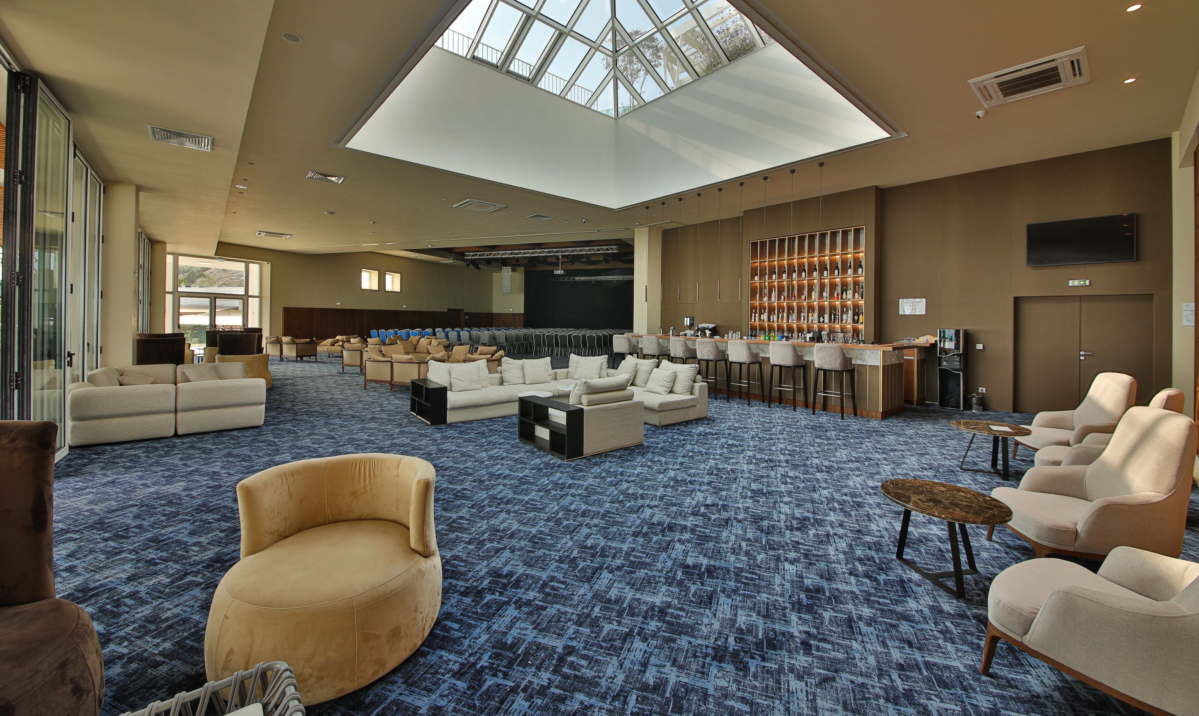 A spacious lounge area with a high ceiling and skylight, seating arrangements, a bar, and a projection screen.