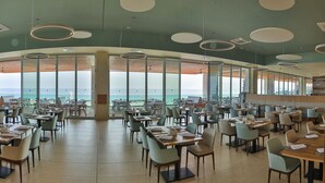 A spacious dining area with tables and chairs, large windows offering a view of the sea, and modern circular ceiling lights.