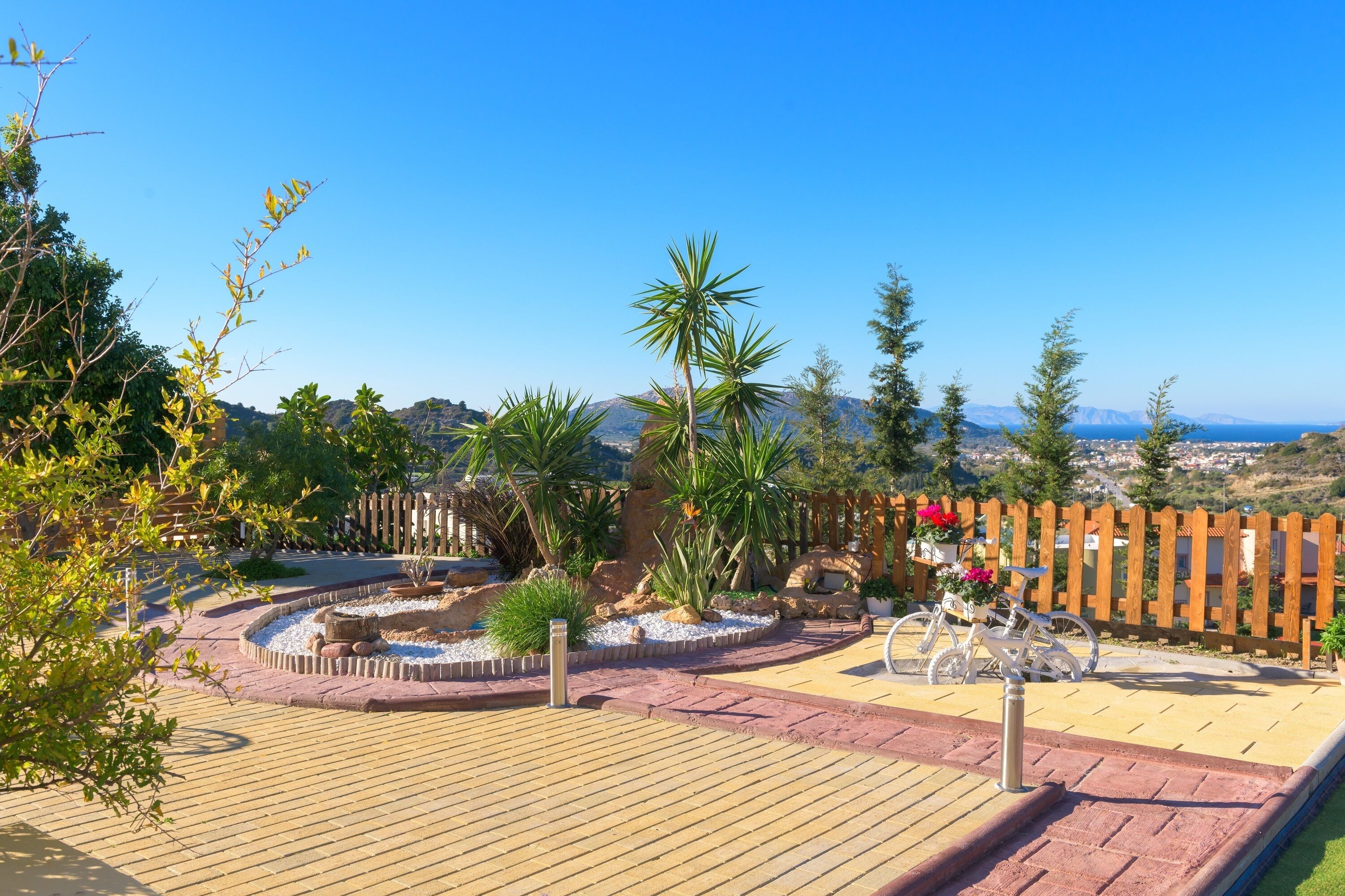 Un jardín ajardinado con una terraza de madera, una fuente y vistas a montañas distantes y un pueblo.