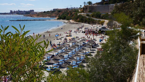 Una playa con numerosas sombrillas y tumbonas, una orilla arenosa y vistas al mar y edificios distantes.