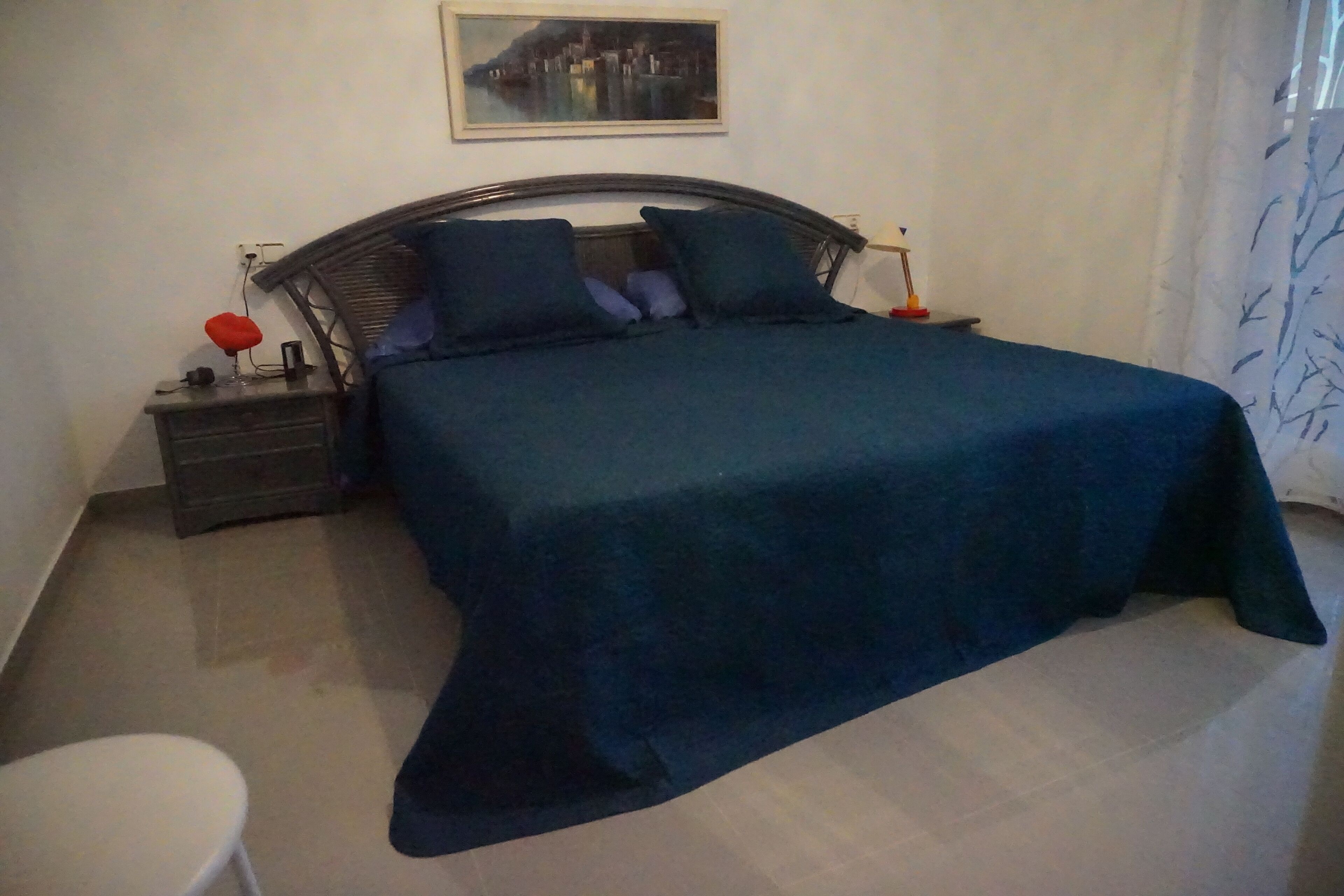 Un dormitorio con una cama, mesitas de noche, una lámpara roja, una silla blanca y un cuadro en la pared.
