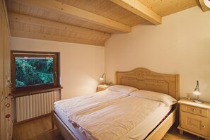 Une chambre avec un plafond en bois, un lit recouvert de draps blancs, une table de chevet avec une lampe et une fenêtre donnant sur un paysage verdoyant.