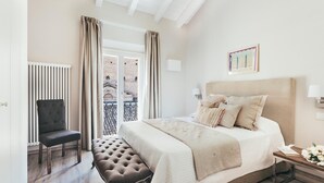 Un dormitorio con una cama grande, un banco, una silla, una mesita de noche y vistas a un edificio a través de la ventana.