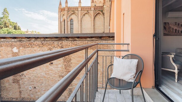 Un balcón con barandilla de hierro forjado, una silla de mimbre con cojín blanco y vistas a una iglesia histórica con campanario.