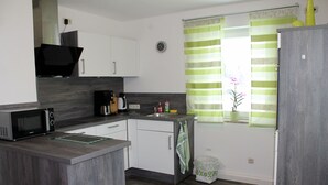 Eine kompakte Küche mit Mikrowelle, Kaffeemaschine und einem kleinen Spülbecken. Ein Mülleimer und ein Fenster mit gestreifter Gardine sind ebenfalls vorhanden.