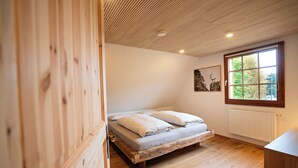 Une chambre avec un plancher en bois, un lit recouvert de draps blancs, une fenêtre donnant sur des arbres et un radiateur.