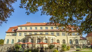 A large, historic building with a curved facade, multiple windows, and a red-tiled roof. There are outdoor seating areas with umbrellas and a large urn in the foreground. - Kurhotel Fürstenhof (Blankenburg)