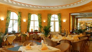 A dining area with wicker furniture, tables set with yellow tablecloths, and large windows with curtains. - Kurhotel Fürstenhof (Blankenburg)