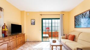 Un salon comprenant un canapé, un téléviseur posé sur un meuble en bois, une table avec un panier, et une porte donnant sur un balcon avec vue sur les arbres.