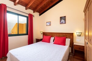 Un dormitorio con cabecera de madera, cama con ropa blanca y almohadas rojas, mesita de noche con lámpara y ventana con cortinas rojas.