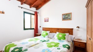 Une chambre à coucher avec une tête de lit en bois, un lit recouvert d’une literie à motifs floraux, une table de chevet avec un livre, et une fenêtre avec des rideaux.