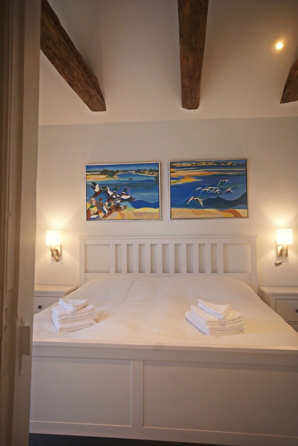 A neatly made bed with folded towels, a white headboard, and two wall-mounted paintings depicting seascapes.