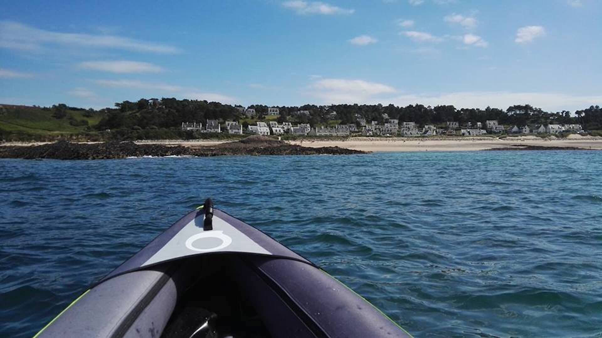Un kayak sur une étendue d’eau calme, avec une plage et des maisons en arrière-plan.