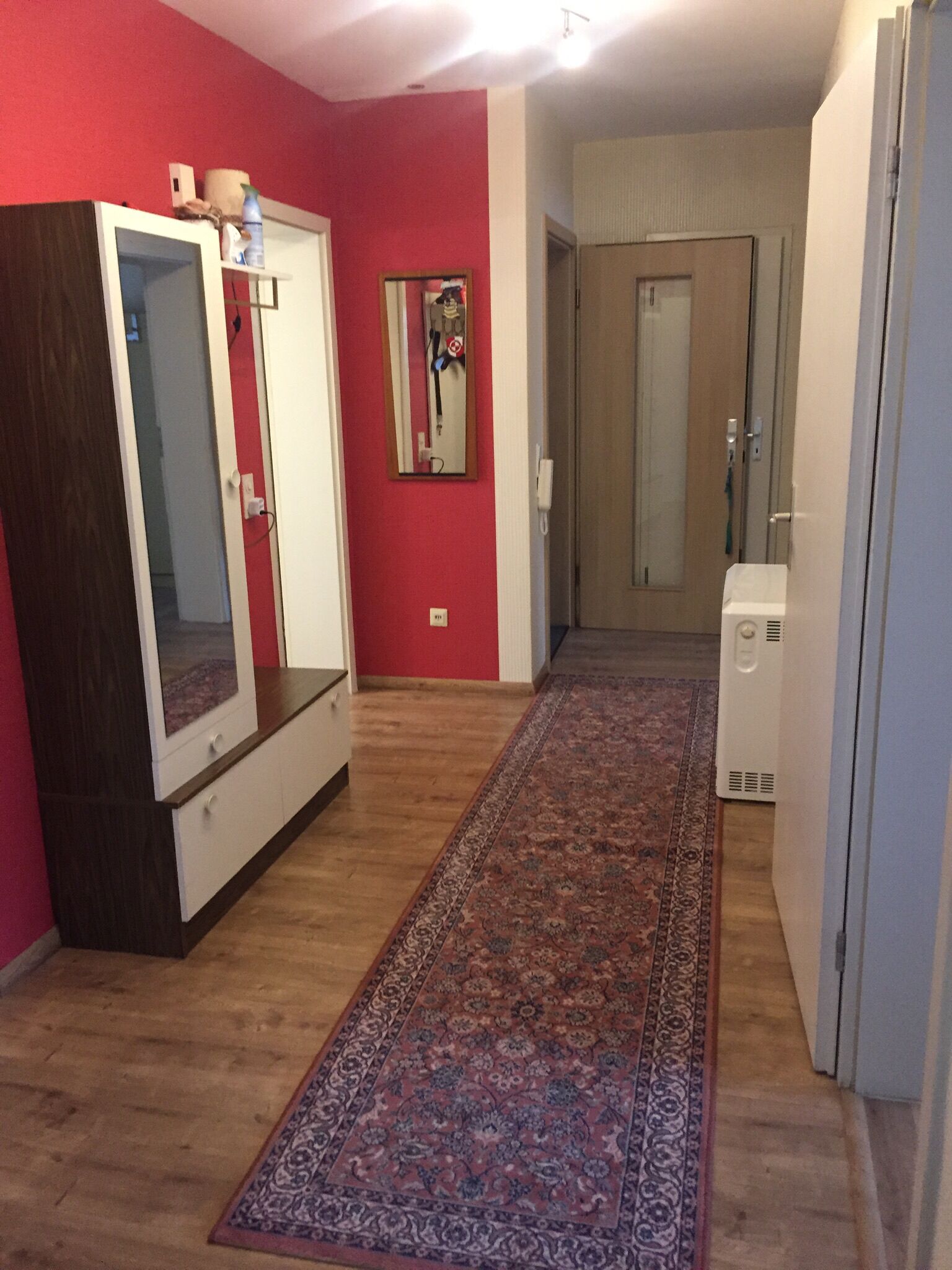 En hall med trägolv, en röd vägg, en vit garderob, en spegel och en dörr som leder till ett annat rum.