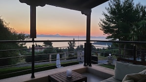 En balkon med udsigt til en solnedgang over vand, omgivet af træer og et fjerntliggende bjerglandskab.