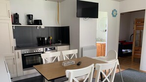Une cuisine compacte équipée d’une cuisinière, d’un four et d’une machine à café, une table à manger avec des chaises, et un téléviseur fixé au mur.