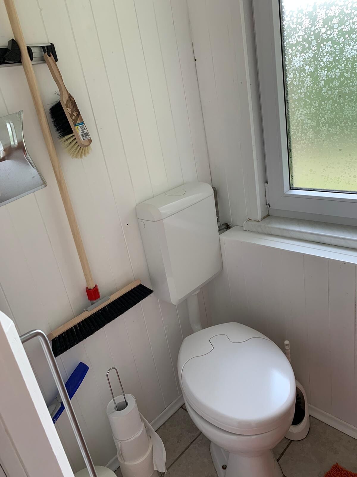 Une petite salle de bain propre, avec des toilettes, une fenêtre et un balai appuyé contre le mur.