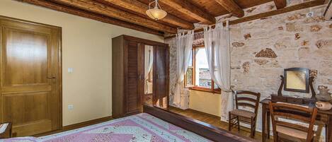 A room with a large bed, wooden ceiling beams, stone walls, a wooden door, and a window with curtains.