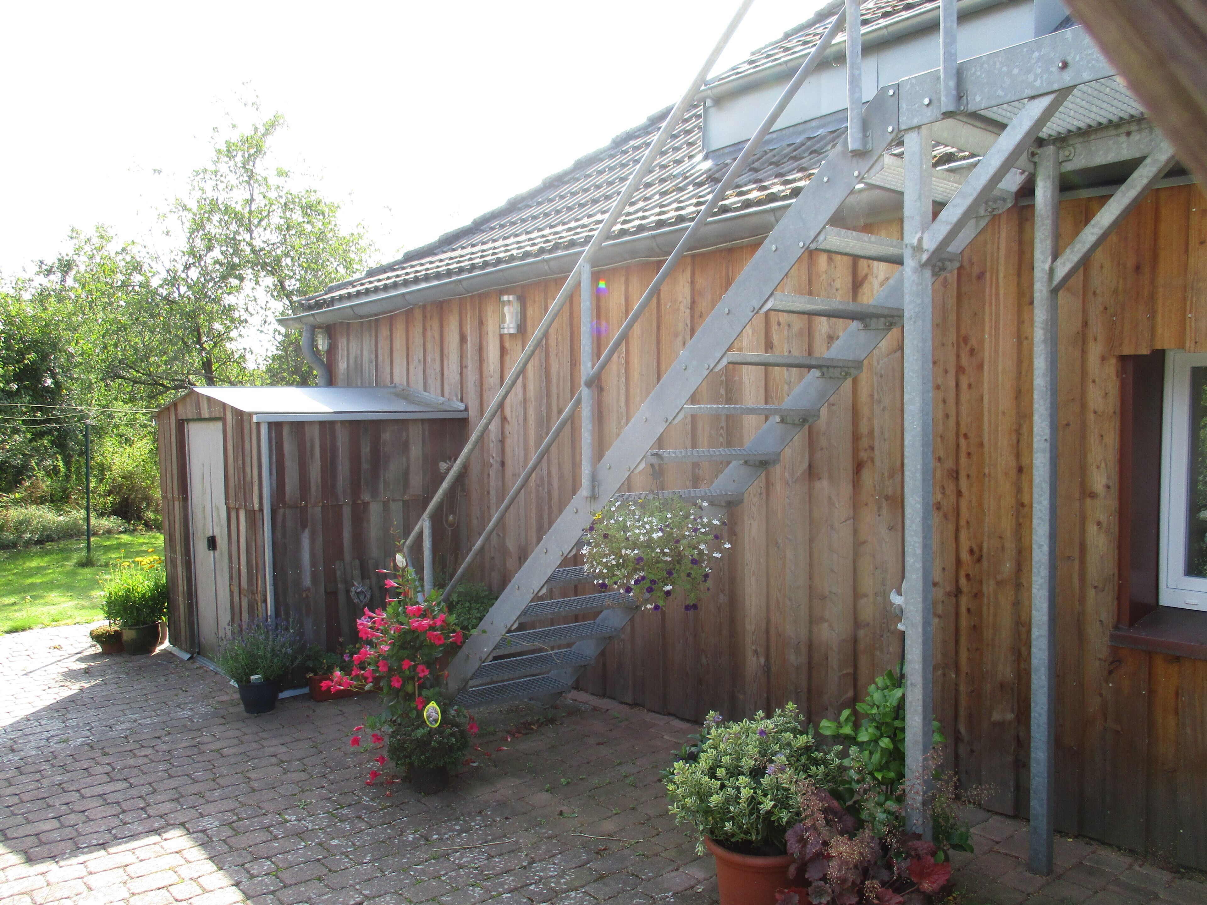 Een houten gebouw met een metalen trap die naar een zolderruimte leidt. Op de voorgrond staat een kleine schuur en staan planten in potten.