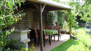 A covered patio with a table and chairs, surrounded by lush greenery and a brick pathway.