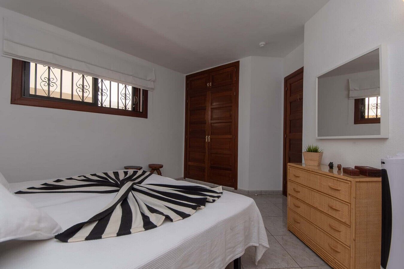 Ein Schlafzimmer mit einem Bett, einem Holzschrank, einem Schminktisch und einem Fenster mit Metallgitter.