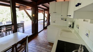 Une cuisine moderne avec des meubles blancs, une cuisinière noire et une machine à café. On y trouve un coin repas avec des chaises et une table en bois, ainsi qu’une terrasse avec une table et des chaises visibles à travers des portes vitrées.