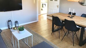 A dining area with a wooden table, black chairs, a white side table with a plant, and a TV mounted on the wall.