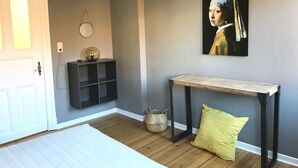 A room with a wooden floor, a white door, a black console table, a yellow cushion, a woven basket, and a painting on the wall.
