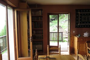 A room with a wooden bookshelf, a dining table with chairs, a window with a view of a garden, and a wall-mounted quilt.