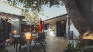An outdoor patio with a table and chairs, a hanging red umbrella, and a large tree providing shade. - BUNGALOW T3 PITAYA (Saint-Paul)