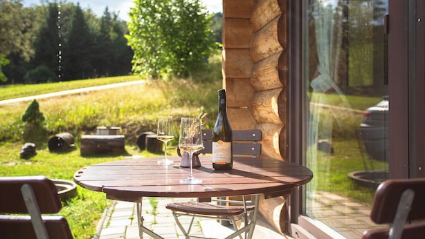 Una mesa de madera con una botella de vino y una copa, ubicada en un patio con vista a un jardín y un edificio.
