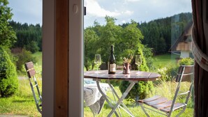 Una mesa con una botella y copas, una silla, y una vista de un paisaje verde a través de una ventana.
