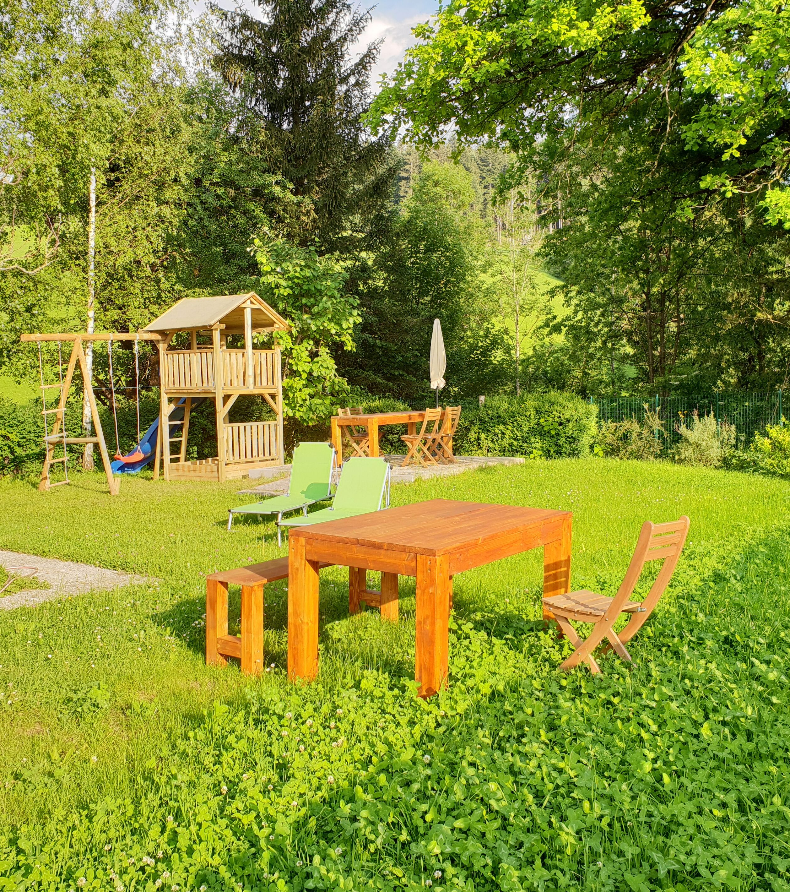 A wooden playhouse with a slide, a picnic table, and two chairs in a grassy backyard.