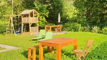 A wooden playhouse with a slide, a picnic table, and two chairs in a grassy backyard.