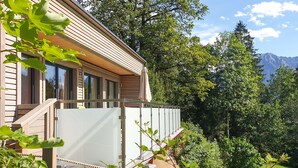 Une maison moderne dotée de grandes fenêtres, d’un balcon et offrant une vue sur une forêt et des montagnes.