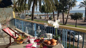 Un balcon avec une table dressée pour le petit-déjeuner, donnant sur une zone côtière bordée de palmiers et offrant une vue sur la mer.