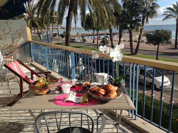 Un balcón con una mesa lista para el desayuno, con vista a la costa, palmeras y el mar.