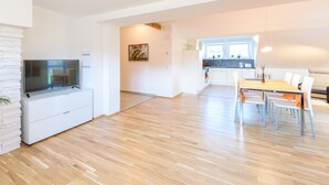 A modern living room with a flat-screen TV, a dining area with a table and chairs, and a kitchen with a window.