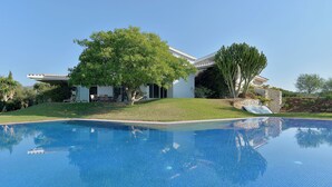 A modern house with a swimming pool, surrounded by greenery and a clear blue sky.