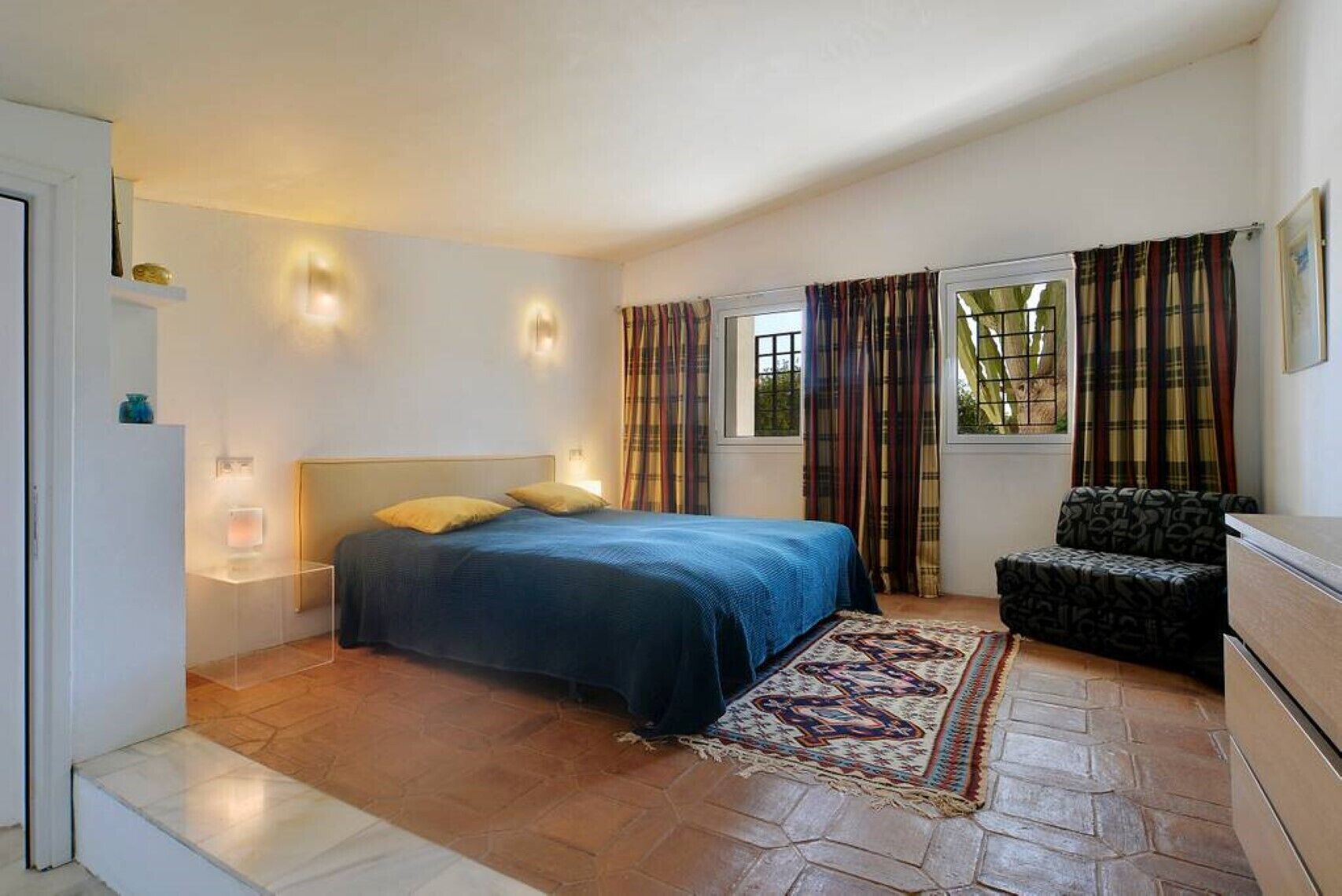 Une chambre à coucher avec un lit, une chaise, une commode et une fenêtre avec des rideaux.
