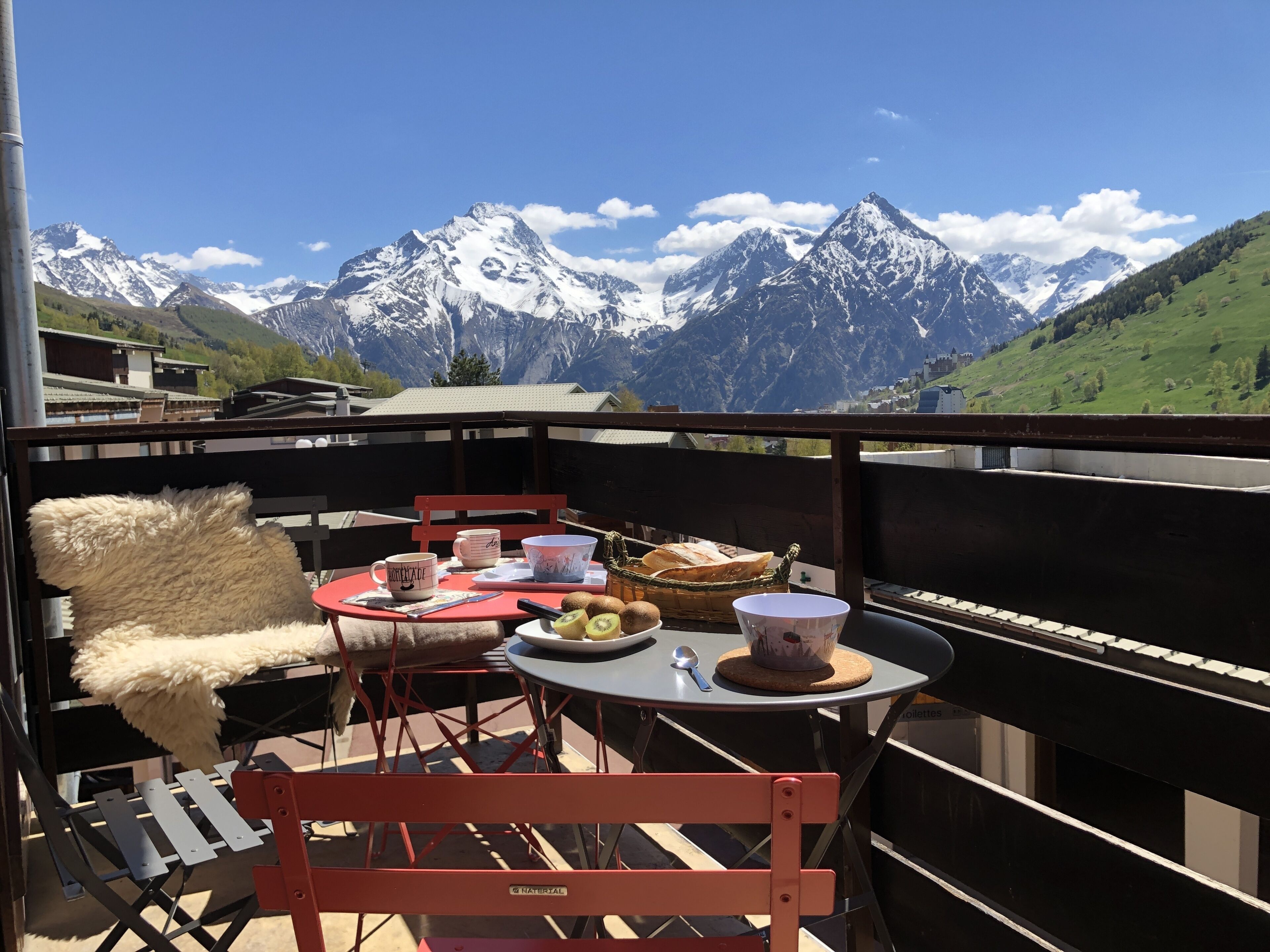 Un balcon avec une table dressée pour un repas, comprenant une chaise, un panier de pain et une vue panoramique sur les montagnes.
