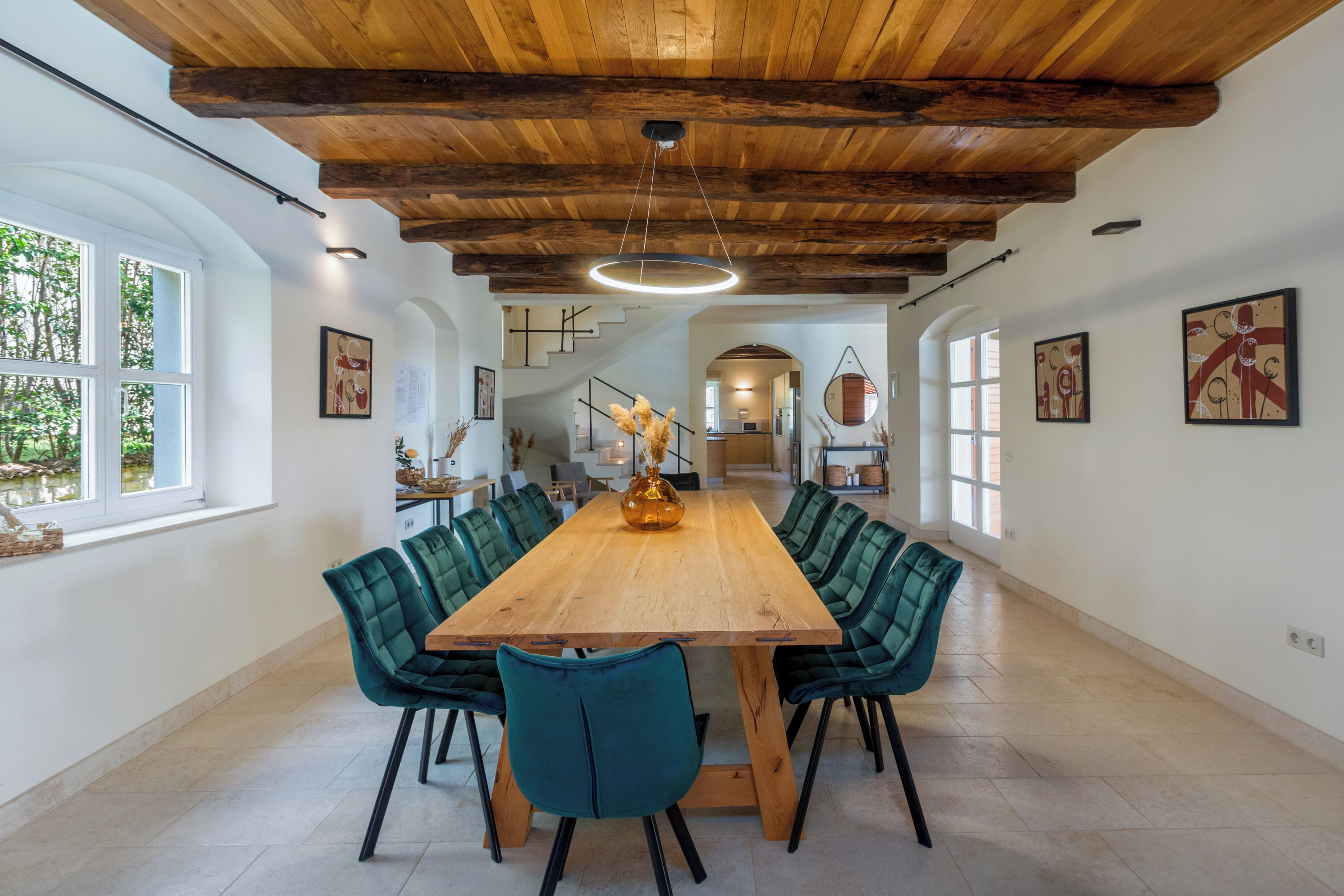 A spacious dining area with a wooden table and teal chairs, a wooden ceiling, and a view of a staircase.