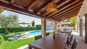 Eine Terrasse im Freien mit einem Holztisch und -stühlen, einer hängenden Leuchte und Blick auf einen Pool und Grünflächen.