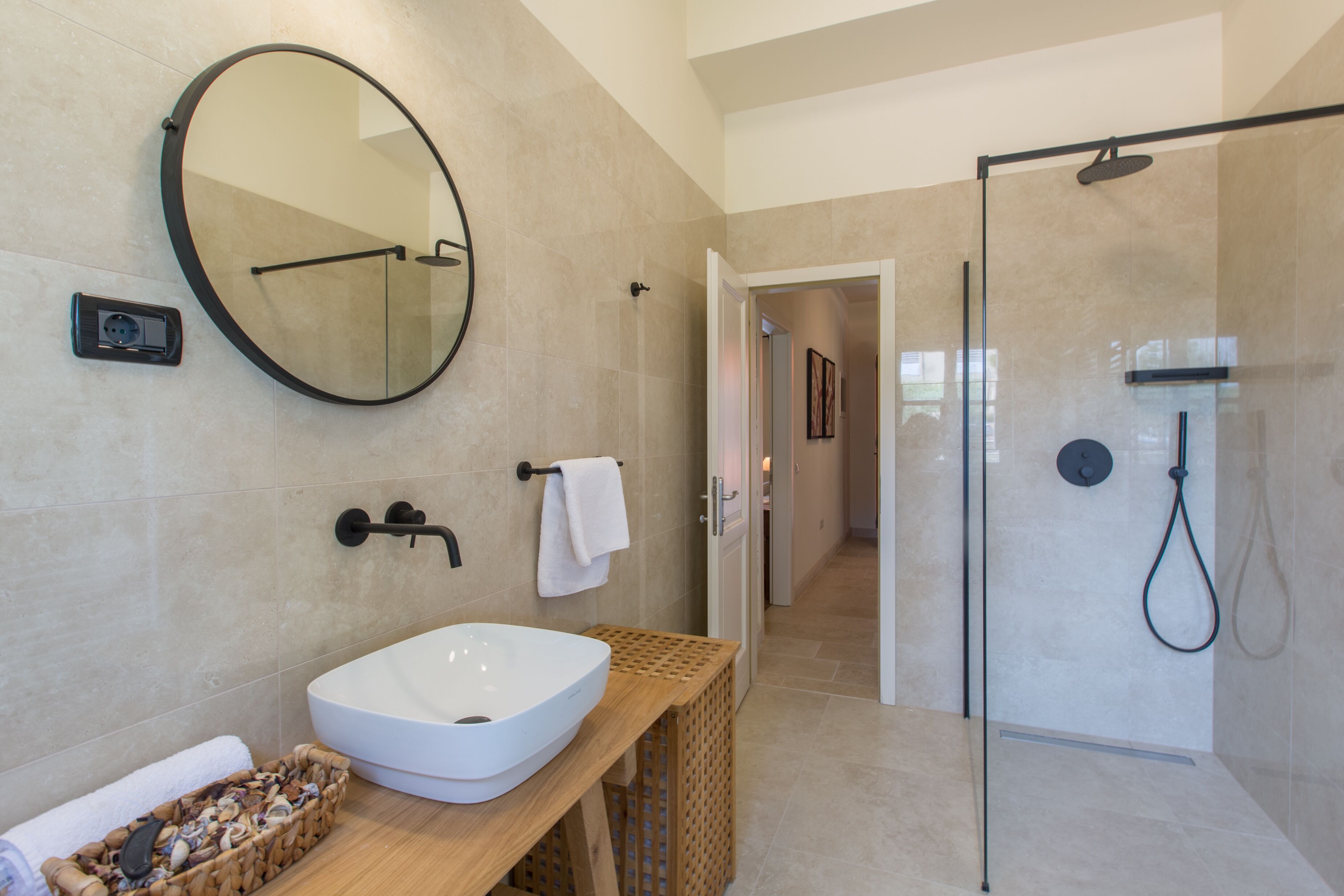 Un baño moderno con un espejo redondo grande, un lavabo blanco, un mueble de tocador de madera y una zona de ducha con mampara de cristal.