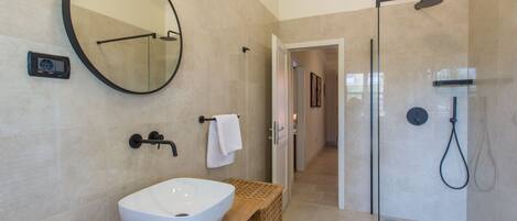 Un baño moderno con un espejo redondo grande, un lavabo blanco, un mueble de tocador de madera y una zona de ducha con mampara de cristal.