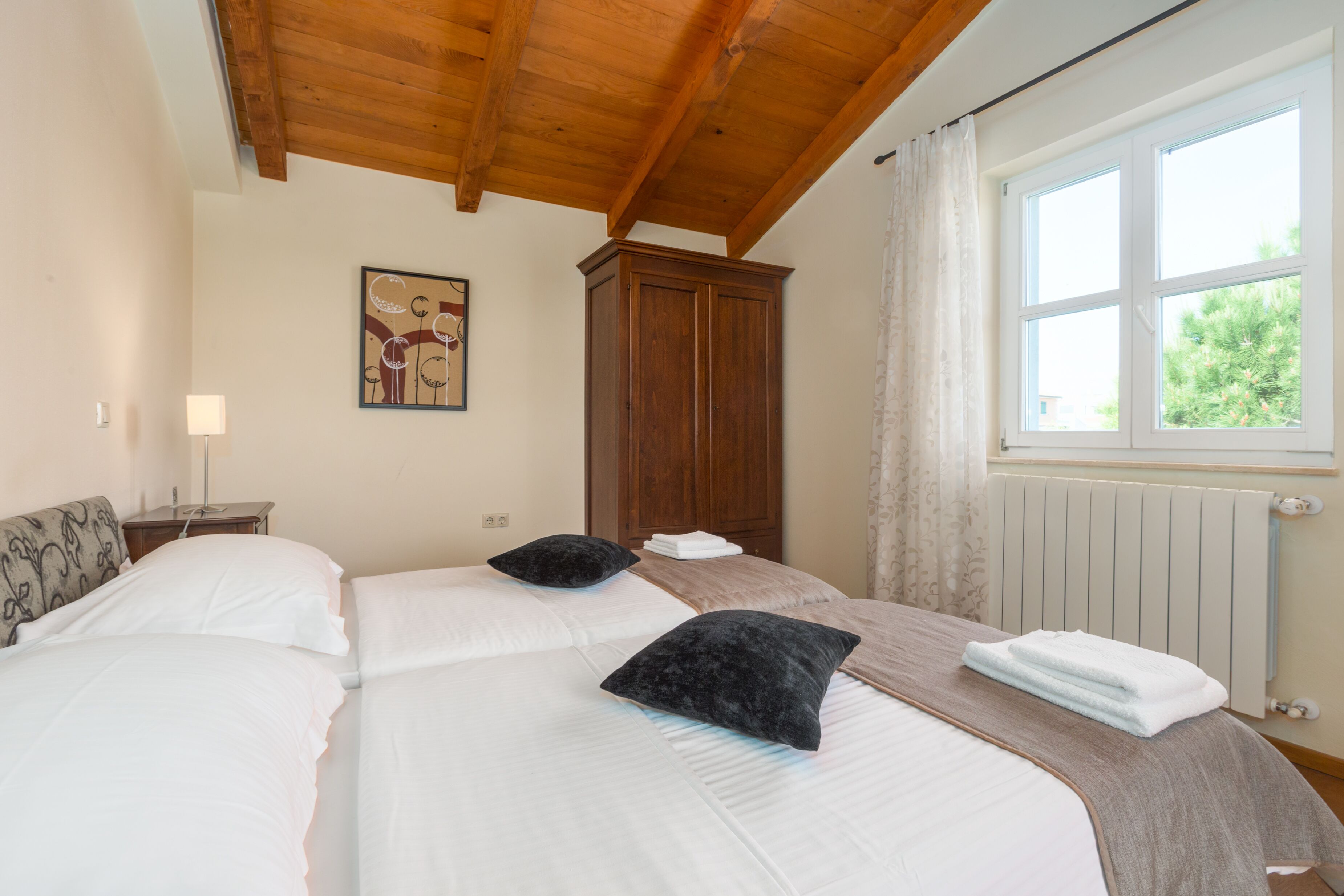 A hotel room with two beds, a wooden ceiling, a window with a view, a radiator, and a cabinet.
