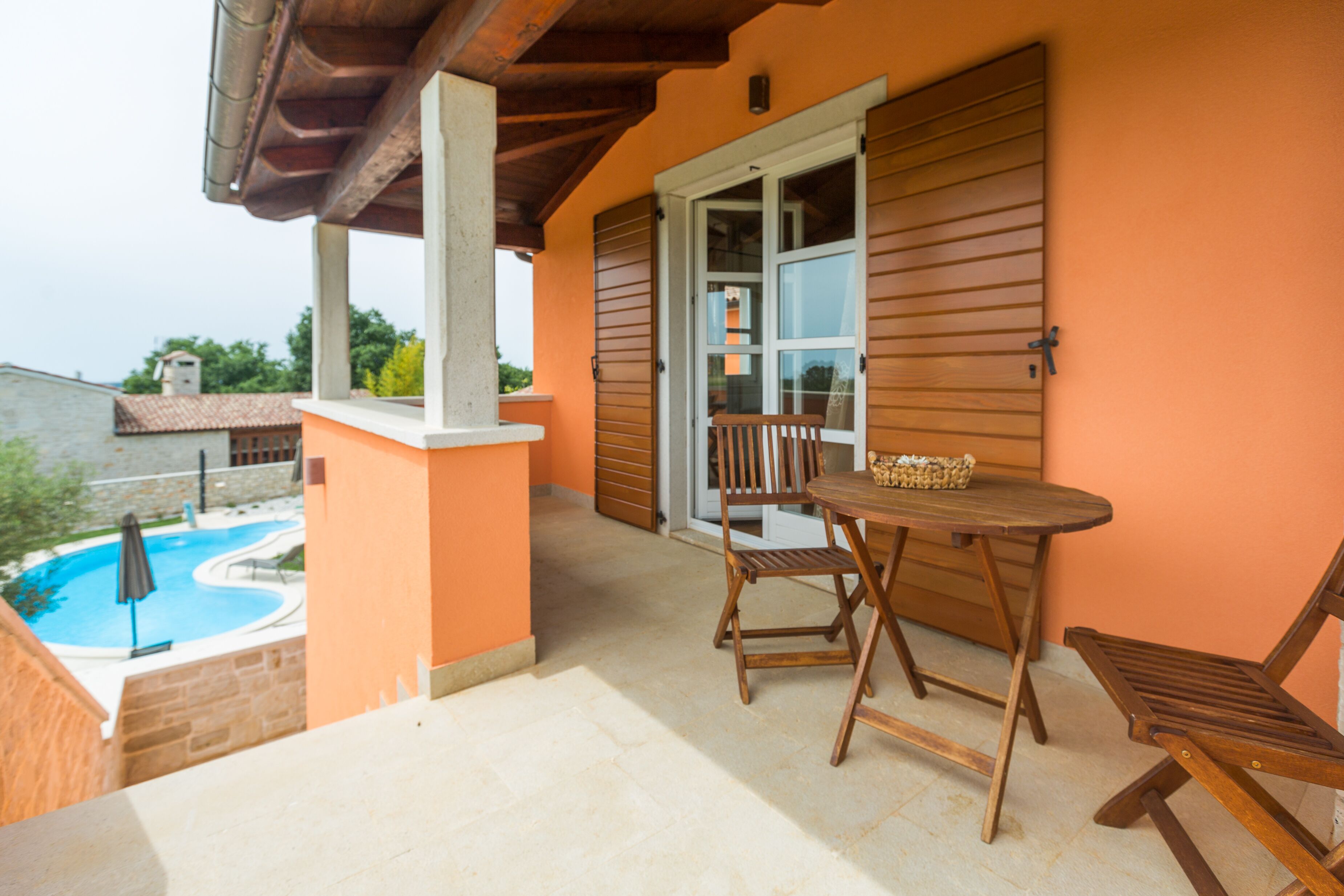 Un balcon avec une table et des chaises en bois, une vue sur une piscine et une porte avec des volets en bois.