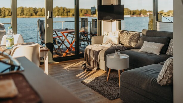 A modern living room with a sofa, coffee table, and a TV on a stand. There is a large window with a view of a lake and a dock.